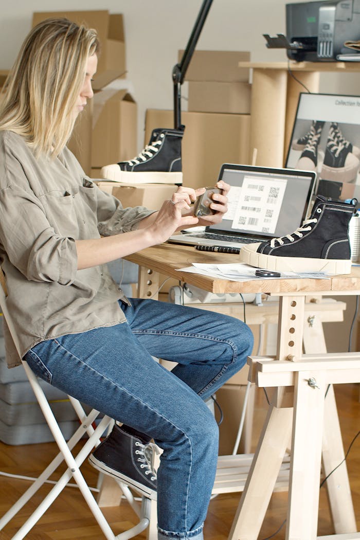 about-02 Woman using laptop and smartphone to manage an online shoe store in a workspace filled with boxes.