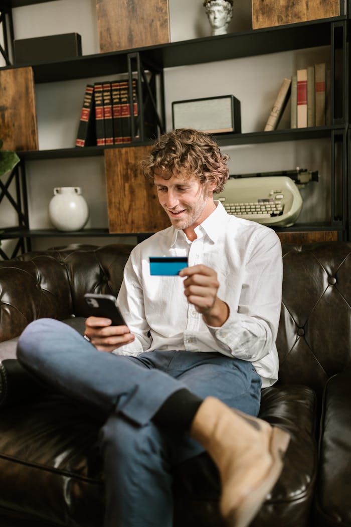 An adult man using a credit card and smartphone for online shopping while sitting on a leather sofa indoors.